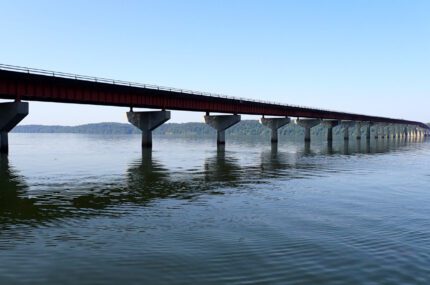 A long bridge with concrete supports across a wide body of water, with distant hills visible under a clear blue sky.