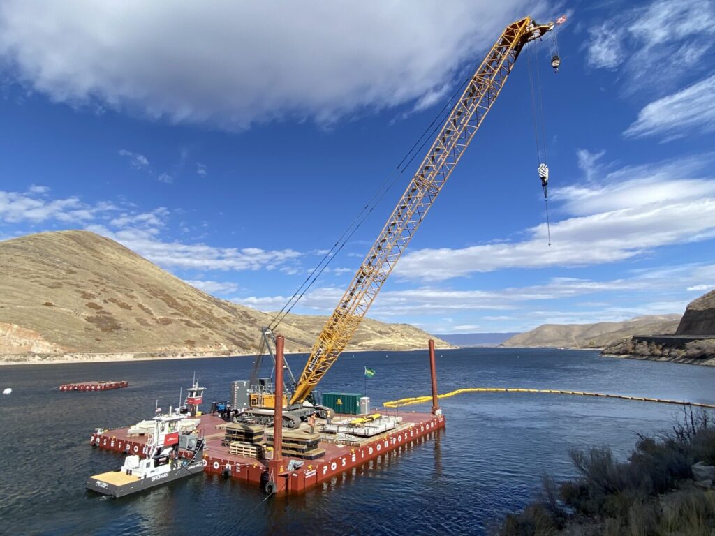 A large crane on a floating barge is positioned on a river surrounded by hills under a partly cloudy sky.