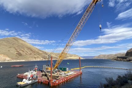A large crane on a floating barge is positioned on a river surrounded by hills under a partly cloudy sky.