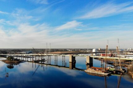 A partially constructed bridge spans a calm river, with cranes and construction equipment visible on both sides under a blue sky with light clouds.