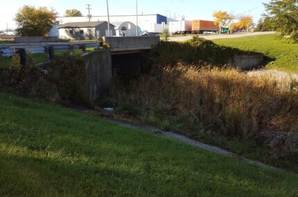 A concrete bridge crosses over a grassy and overgrown drainage ditch near an industrial area with warehouses and parked semi-trailers in the background.