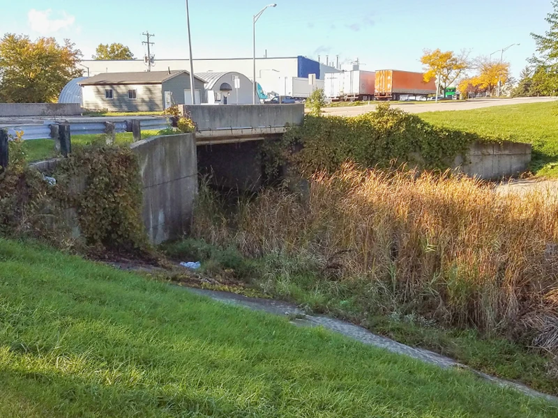 A concrete culvert overgrown with vegetation runs beneath a road near an industrial area with buildings, parked trucks, and autumn trees in the background.