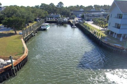 A small marina with a docked boat, wooden piers, parked cars, and buildings surrounded by trees on a sunny day.