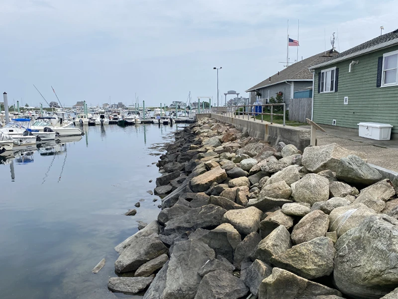 A marina with docked boats, a rock-lined shoreline, and a green building with an American flag on a cloudy day.