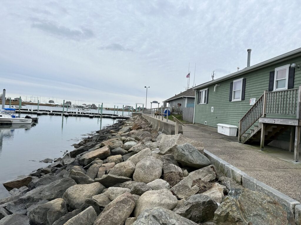 A green building with stairs stands by a rocky shoreline next to a marina with boats and docks under a cloudy sky.