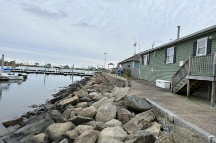 A green building with stairs stands by a rocky shoreline next to a marina with boats and docks under a cloudy sky.