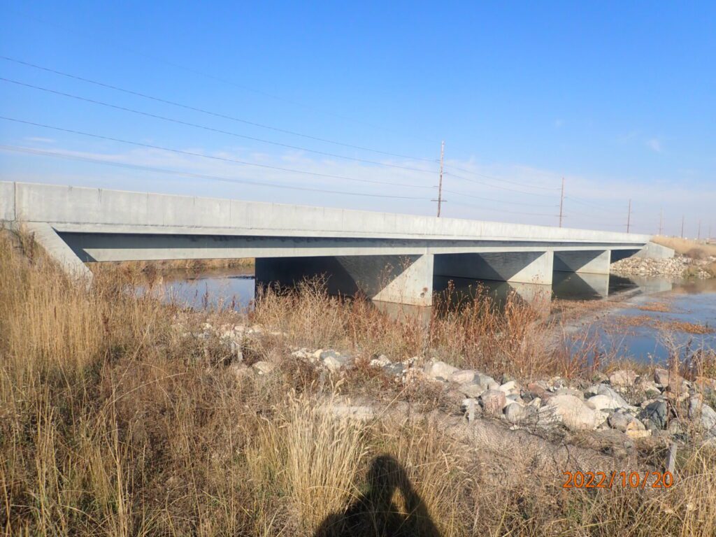 A concrete bridge crosses a shallow body of water with dry grass and rocks in the foreground; utility poles are visible in the background.