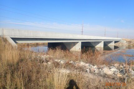 A concrete bridge crosses a shallow body of water with dry grass and rocks in the foreground; utility poles are visible in the background.
