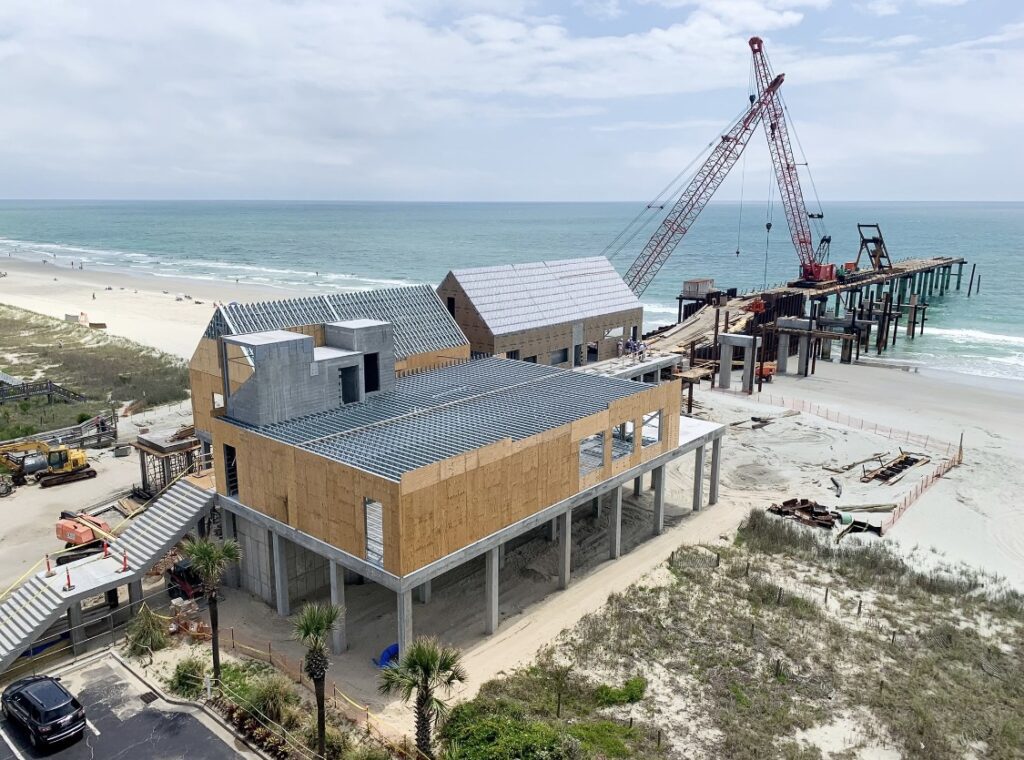 A coastal construction site with a large building on stilts and a crane working on a pier extending into the ocean.