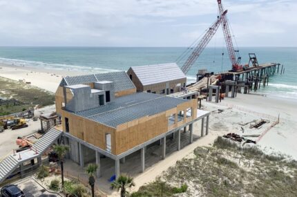 A coastal construction site with a large building on stilts and a crane working on a pier extending into the ocean.