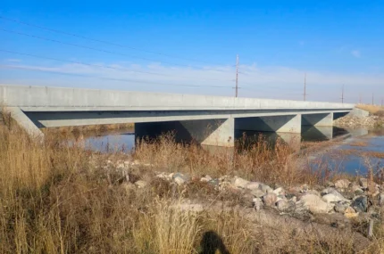 A concrete bridge spans over a shallow body of water, surrounded by dry grasses and rocks under a clear blue sky.