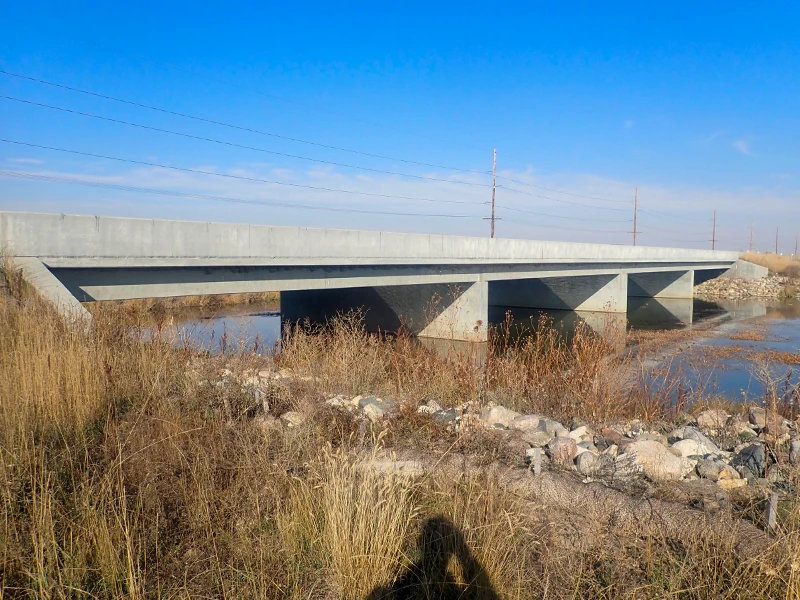 A concrete bridge spans over a shallow body of water, surrounded by dry grasses and rocks under a clear blue sky.