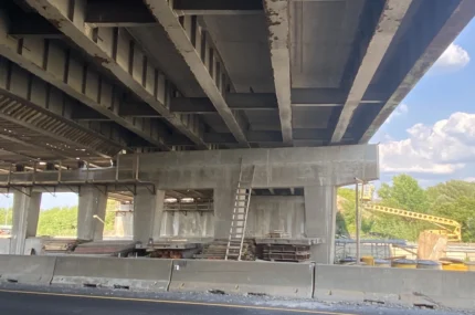 View under a highway overpass with construction materials, a ladder leaning on concrete supports, and a crane visible in the background.