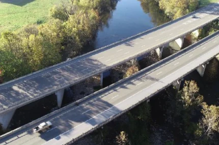Aerial view of two parallel highways crossing over a river, surrounded by trees and grassy areas, with a vehicle on one road.