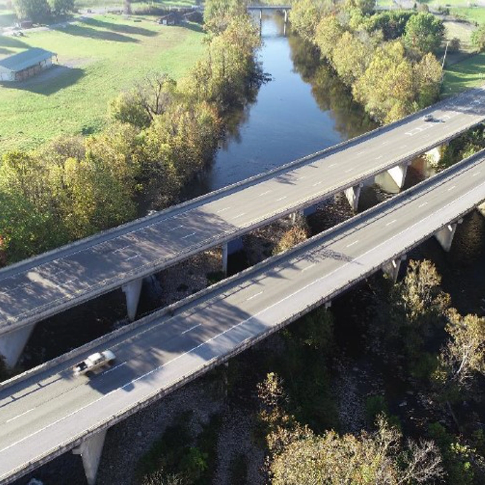 Aerial view of two parallel highways crossing over a river, surrounded by trees and grassy areas, with a vehicle on one road.