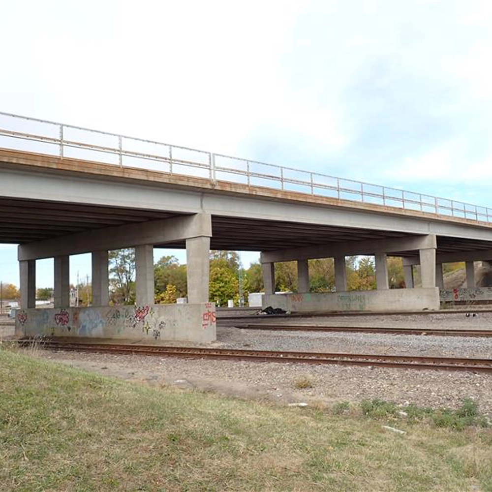 A concrete overpass crosses above railway tracks, with graffiti visible on the bridge supports and grass in the foreground.