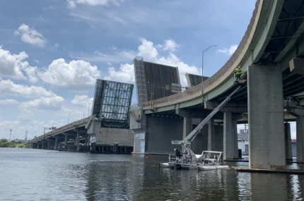 A drawbridge over a river is raised. Workers on a platform perform maintenance on the bridge structure. The sky is partly cloudy.