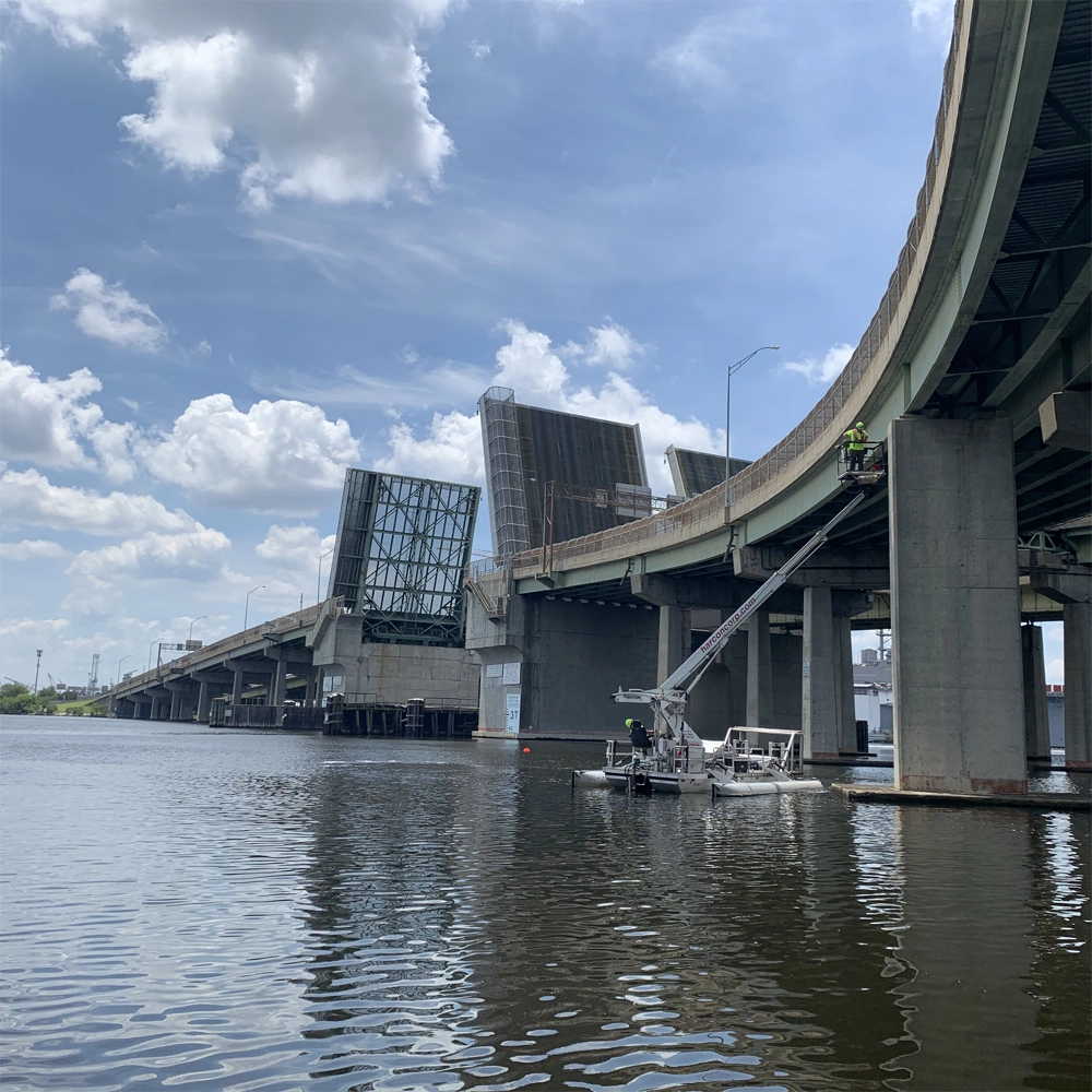 A drawbridge over a river is raised. Workers on a platform perform maintenance on the bridge structure. The sky is partly cloudy.