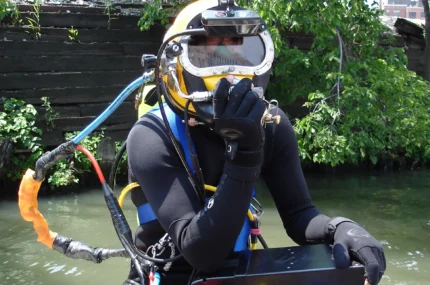 Person in wetsuit and full-face diving mask sits beside water, holding a communication device and equipment box, with greenery and a wooden structure in the background.