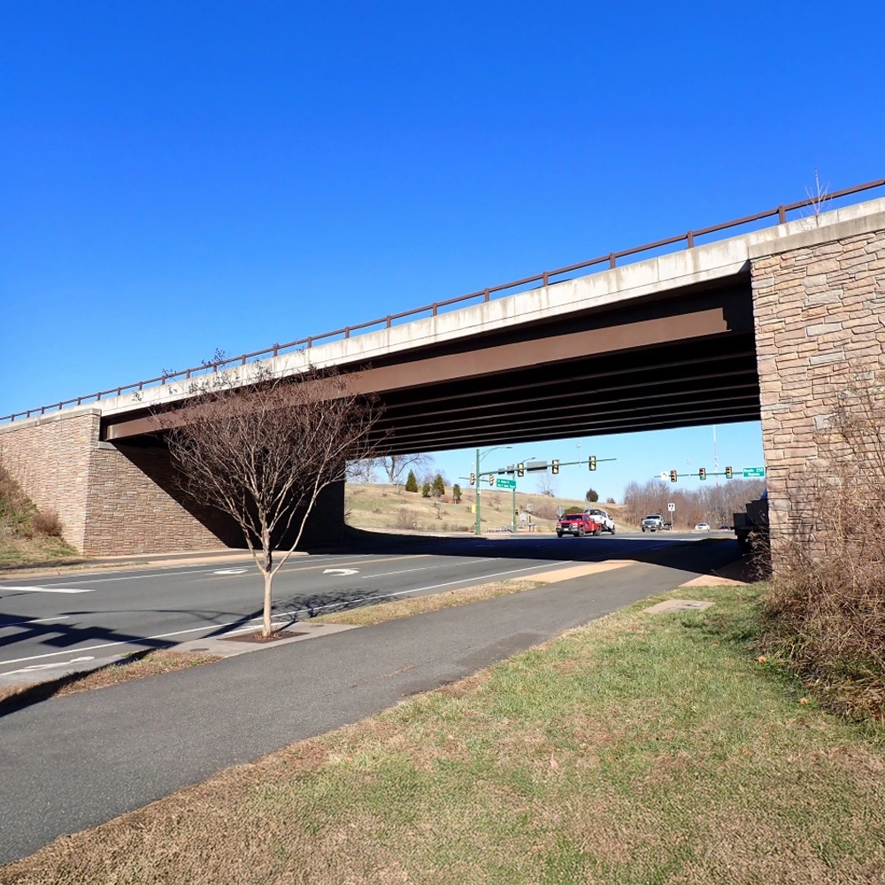 A concrete and stone bridge crosses over a road with a leafless tree nearby; cars and traffic lights are visible in the background under a clear blue sky.