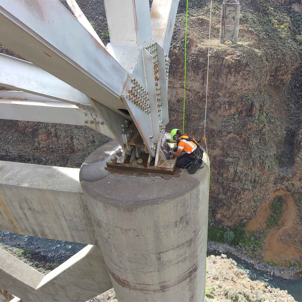 Worker in safety gear inspects or repairs a bridge support high above a canyon, with rocky cliffs and a river visible below.