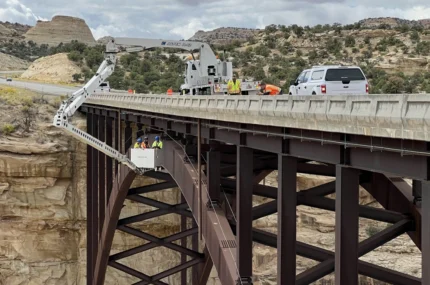 Workers in a bucket are suspended below a steel bridge by a specialized truck, performing maintenance or inspection; a white vehicle and equipment are on the roadway above.