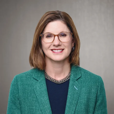 A woman with shoulder-length brown hair, wearing glasses, a green blazer, a dark top, and a chunky necklace, smiles in front of a neutral background.
